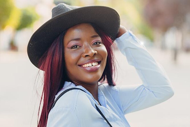 Portrait einer jungen, lachenden afroamerikanischen Frau mit pinken Haaren, die einen Hut träg