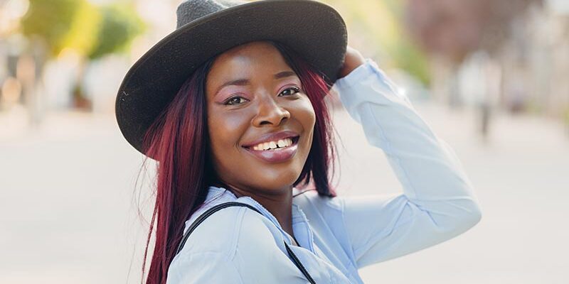 Portrait einer jungen, lachenden afroamerikanischen Frau mit pinken Haaren, die einen Hut träg Portrait einer jungen, lachenden afroamerikanischen Frau mit pinken Haaren, die einen Hut träg